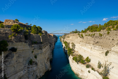 Clear blue water flows through the narrow Corinth Canal, bordered by steep, sunlit limestone walls topped with green pines. An arched bridge spans the dramatic gorge beneath a vibrant sky, creating a