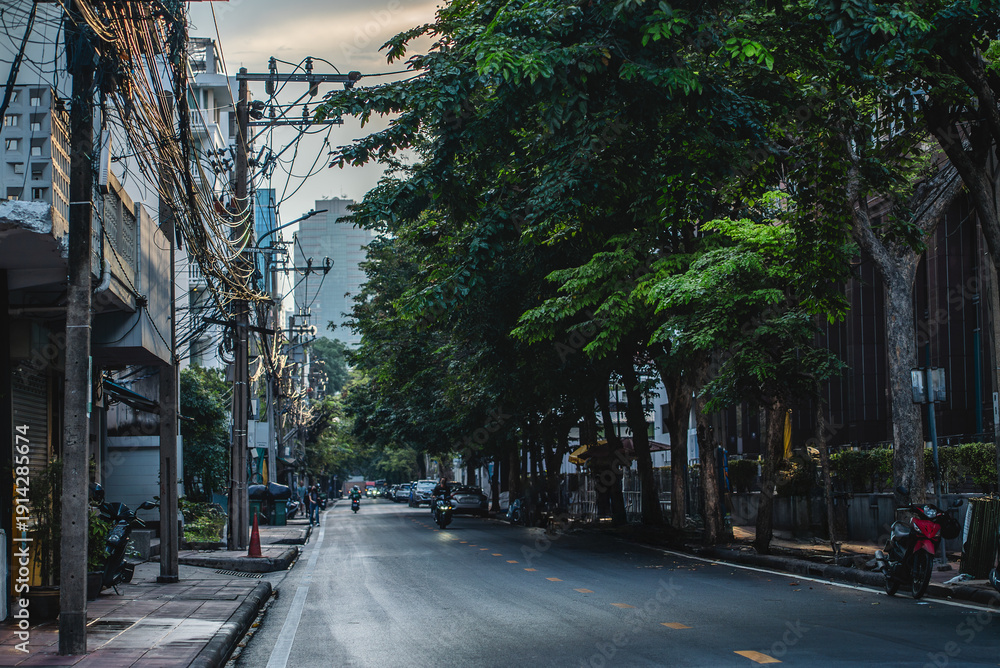 Obraz premium Typical Bangkok street at sunset with tropical trees and power lines, Cozy urban road in Thai city at dusk with greenery, motorcycles and cables overhead, Thailand