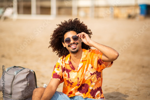Latino man smiling and adjusting sunglasses while sitting on beach