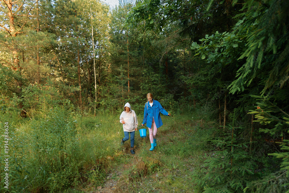 Fototapeta premium Two people walk through a forest path carrying a bucket during a sunny day in the woods