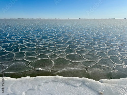 Fotografía Polygonal ice formations on frozen Baltic Sea surface near snowy shoreline, natural winter patterns created by freezing water under clear sky