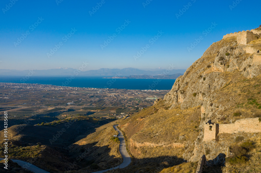 Fototapeta premium Golden rocky slopes of Acrocorinth with ancient stone walls descend toward a vast plain and the shimmering Corinthian Gulf under a clear blue sky in Greece.