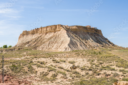 Steep beige cliffs rise from a semi-arid plain, topped by a flat plateau and scattered structures in Bardenas Reales, Spain. Sunlight highlights rugged textures and dry grasses under a clear blue sky.