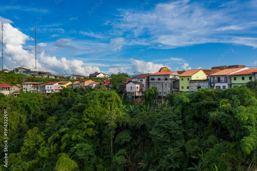 Fototapeta premium Cluster of vibrant houses perched on a lush green hillside with dense tropical foliage below, set against a vivid blue sky and scattered clouds in rural Phou Khoun, Laos. The scene highlights the