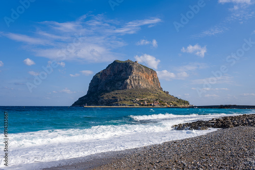 Foto Powerful turquoise waves roll onto a stony beach facing the iconic Monemvasia rock, with scattered clouds and vivid sunlight highlighting the dramatic coastal scenery