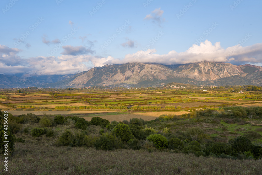 Obraz premium Layered agricultural fields stretch across a broad plain toward rugged Greek mountains partly covered by drifting clouds. Soft afternoon sunlight highlights the varied textures and green patches in
