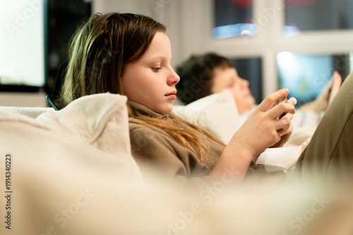 Schoolchildren at a cozy home on a sofa, focused on their smartphones playing mobile games. Scene captures modern childhood and the growing issue of phone addiction, digital dependency