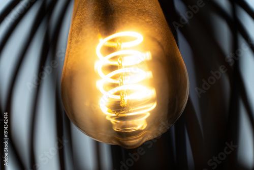 A close-up view of the spiral filament of a glowing bright light bulb inside a lampshade. The intricate details of illuminated coil create a warm and inviting atmosphere.