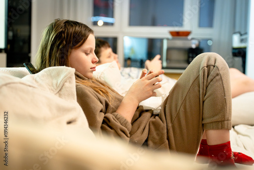 Two children, a boy and a girl, siblings on a sofa at home, deeply focused on a smartphone playing games. Concept of phone addiction, digital dependency