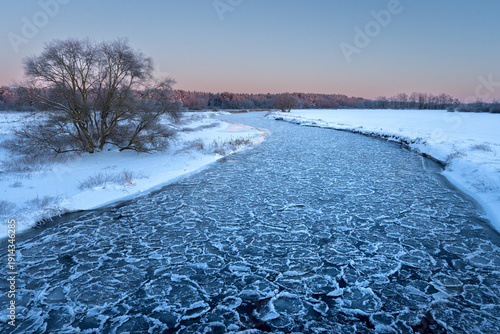 Beautiful nature landscape of winter river with cracked ice at sunset. Warm golden light reflects off the frozen surface, with a dark forest silhouette on the horizon.