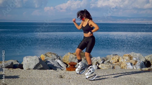 A beautiful sexy girl in black tight sport body perform excerises on kangoo shoes on the pier next to the blue sea with white clouds at the background. Rock'n'roll, boxing and tae-bo moves