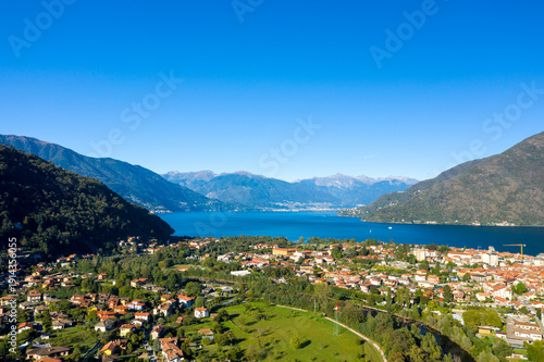 Quadro su tela Aerial panorama of Cannobio on the shore of Lake Maggiore in northern Italy, framed by forested hills and distant alpine peaks beneath a vivid blue sky