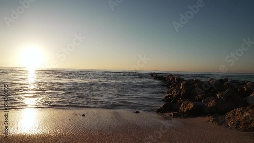 Low-angle sunrise view of sand on a beach, with rocks and waves crashing onto the shore, on a bright, beautiful, blue-sky summer day.