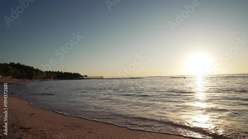 Low-angle sunrise view of sand on a beach, with rocks and waves crashing onto the shore, on a bright, beautiful, blue-sky summer day.