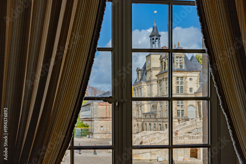 Elegant interior window view with striped curtains overlooking Château de Fontainebleau’s ornate stone facade, tower, and grand staircase under blue sky, UNESCO World Heritage Site, France