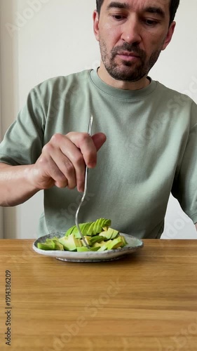Middle aged man enjoying a healthy meal. He is eating fresh sliced avocado with a fork while sitting at a wooden table in his kitchen