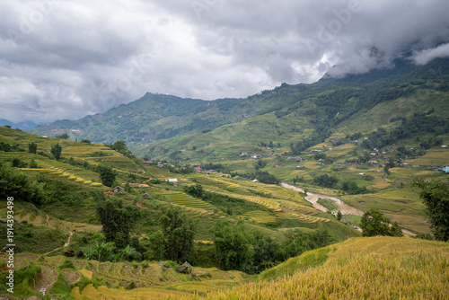 Canvas Print Layered rice terraces sweep across the lush valley near Sapa, with a winding river and scattered rural houses beneath dramatic, cloud-covered mountains