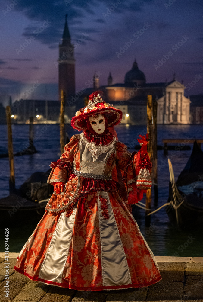 Fototapeta premium Masked woman in ornate orange jester costume, and holding a little lookalike jester doll, poses for the camera at San Marco, at the annual Venice Carnival.
