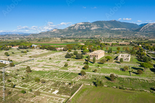 Wide aerial perspective of the ancient archaeological site of Paestum, Italy, featuring expansive stone ruins and two well-preserved Greek temples set amid green fields. The landscape is framed by