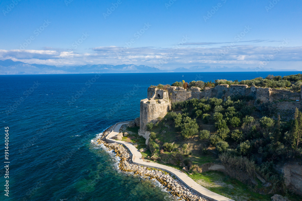 Naklejka premium Ancient stone ramparts overlook the deep blue Mediterranean, bordered by lush olive groves and a winding coastal path under a bright sky near Koroni, Greece.