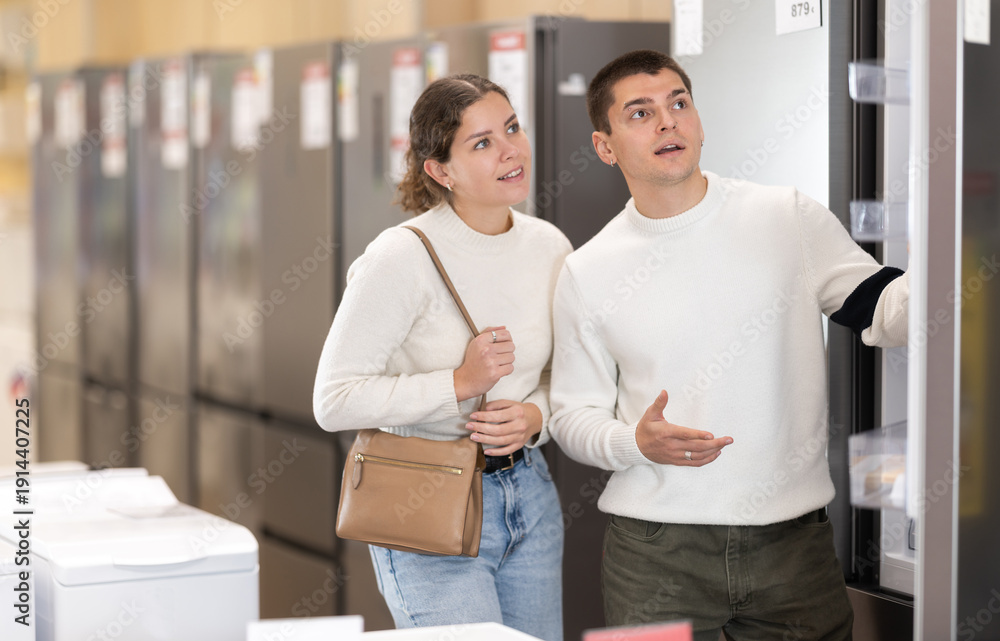 Fototapeta premium Couple customer inspect product range and selects fridge. Client in hardware store picks up household appliances. Store of household appliances and household goods, showroom.