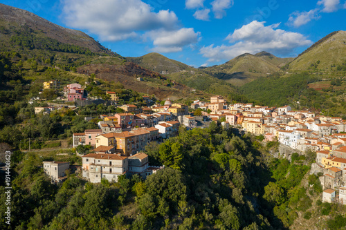 Fotografía Vibrant buildings with terracotta roofs line the steep hillside of Tortora, Italy, surrounded by lush greenery and dramatic mountain peaks under a bright blue sky with scattered clouds