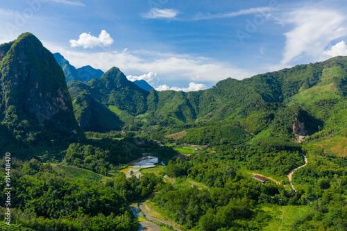 Sweeping view of a lush valley surrounded by dramatic limestone mountains and dense tropical forest near Nong Khiaw, Laos. Bright daylight highlights rice paddies, winding roads, and vibrant
