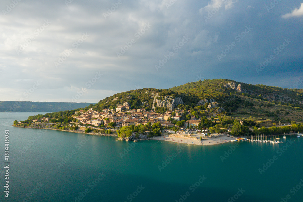 Fototapeta premium Sunlit village of Bauduen sits on a peninsula surrounded by turquoise waters, with a sandy beach and marina nestled beneath green hills and dramatic rocky outcrops under a cloudy sky.