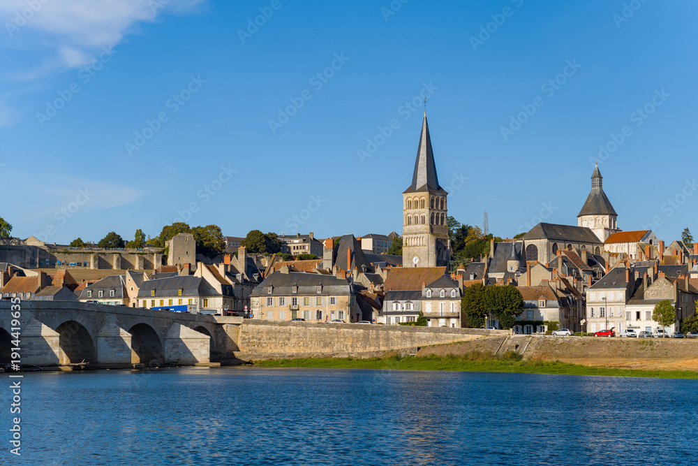 Fototapeta premium Warm evening sunlight highlights the medieval church spires, stone bridge, and traditional rooftops of La Charite sur Loire, with the calm Loire river in the foreground. The scene captures the