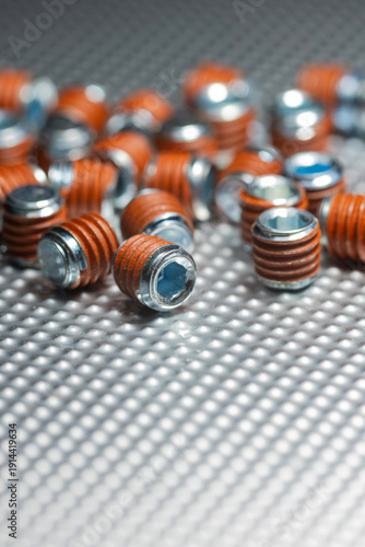 Macro view of industrial grub screws with orange thread locker coating scattered on a textured metallic surface featuring selective focus and technical hardware details