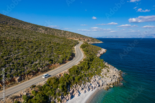 A curving road hugs the rocky coastline of the Peloponnese, bordered by green hills and clear blue water under a sunny sky. The scene conveys a sense of freedom, travel, and natural beauty.