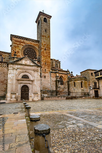 view of the Romanesque and Gothic Cathedral of Santa Maria in Siguenza Spain featuring the stone facade rose window and bell tower over a quiet cobblestone square under a cloudy sky