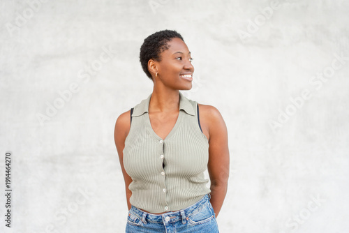 Smiling young woman looking away while standing against gray wall. Thoughtful mood with relaxed posture and modern casual style