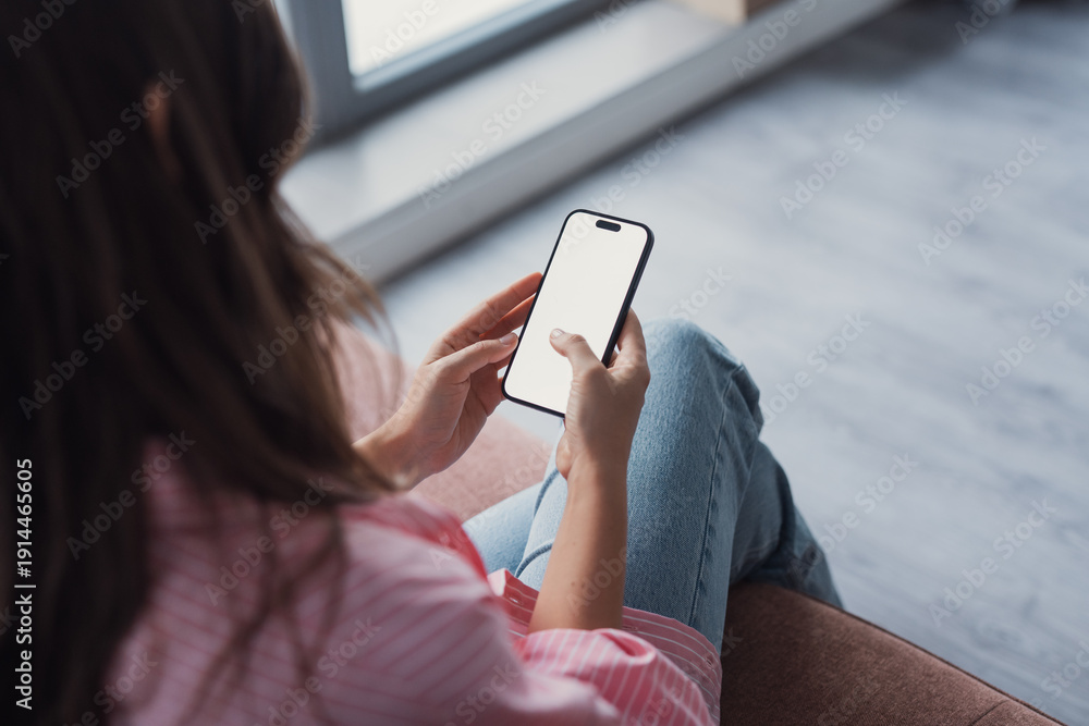 Fototapeta premium Close up shot of young female relaxing indoors at home holding mobile phone with blank screen for mock up in hands, millennial woman scrolling news online, browsing internet sites, calling or chatting