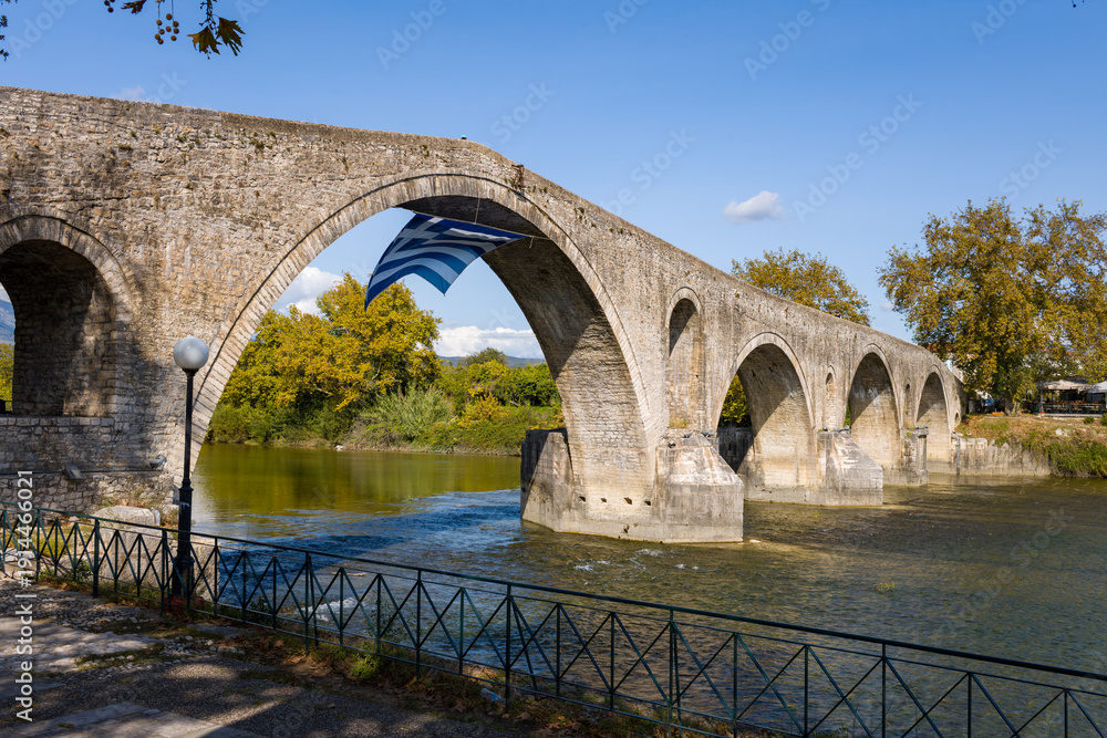 Fototapeta premium Historic multi-arched stone bridge stretches across a calm river in Arta, Greece, with a large Greek flag suspended beneath one of the central arches. Autumn trees and textured masonry are illuminated