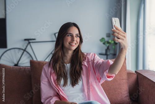 Happy pleasant millennial woman relaxing on comfortable couch, holding smartphone in hands. Smiling young lady taking a selfie, posting on social media, using mobile applications at home.