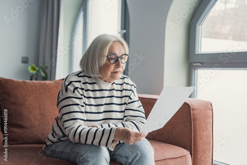 Canvas-taulu Focused elderly 60s woman sit on sofa in living room reading postal corresponden