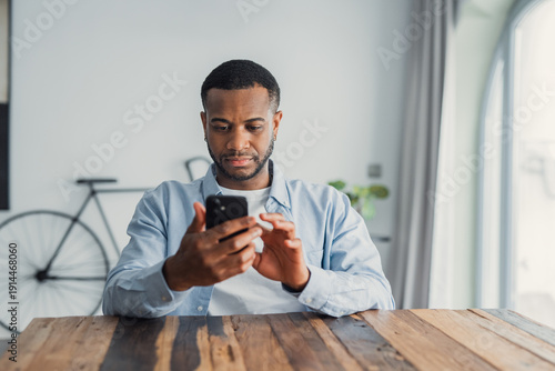 Fotografía Confused African guy holding smart phone feels concerned thinking over received message