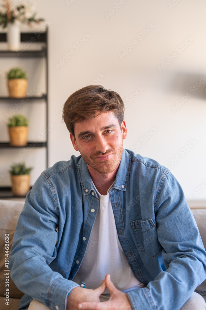 Naklejka premium A cheerful young Caucasian man sits on the couch at home with crossed arms, smiling confidently at the camera and enjoying a relaxed and positive moment in a comfortable indoor setting