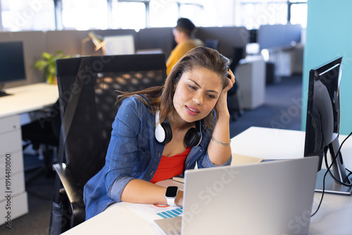 Diverse female coworkers sitting at office desk focusing on laptop with charts, headset, smartwatch
