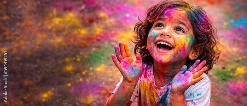 Indian kid smiling with colorful powdered face and hands, celebrating Holi festival. Joyful Hindu spring festival for greeting cards, copy space, banner