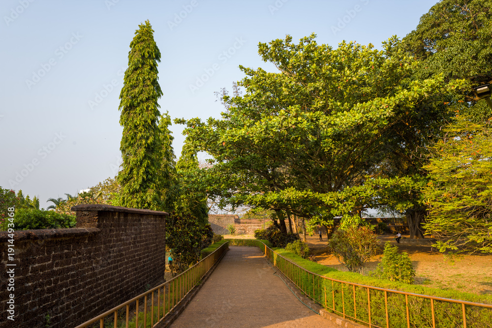 Naklejka premium Tree-lined path runs alongside a historic stone wall at Kannur Fort, India, with lush greenery and warm afternoon sunlight casting soft shadows on the ground. The tranquil scene features textured