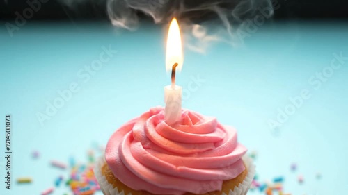 Pink frosted cupcake with lit candle and colorful sprinkles on blue background.