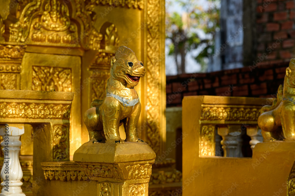Fototapeta premium Close-up of a gold-painted guardian lion sculpture with intricate detailing, set against ornate golden temple architecture at Phnom Sampov in Battambang province, Cambodia. The scene is illuminated by
