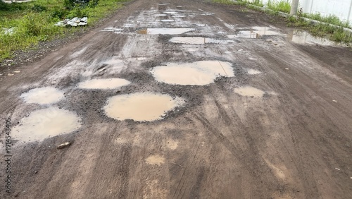 Fotografie Muddy dirt road is filled with many murky water potholes and clear vehicle tire tracks, depicting difficult and neglected unpaved rural infrastructure after rainfall