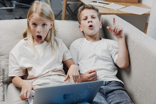Obraz na plátně Two surprised children sit on a sofa using a laptop, reacting with shock and curiosity
