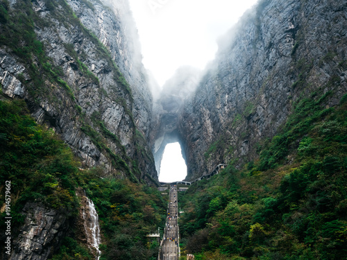 Fotografía Majestic view of Heaven's Gate at Tianmen Mountain in Zhangjiajie, China, featuring dramatic cliffs with fog, lush greenery, and the iconic natural arch surrounded by mist and stone stairs