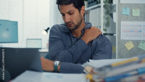 Young Latino businessman suffering shoulder pain and neck ache at office desk with computer. Office syndrome, muscle massage, overworked stress, bad posture, and health insurance injury concept.