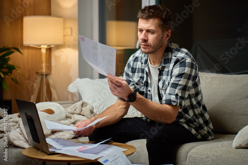Wallpaper Mural Man sitting on couch at home examining paper bills and financial documents with laptop on table. Concept of tracking personal expenses and managing monthly household payments. Torontodigital.ca