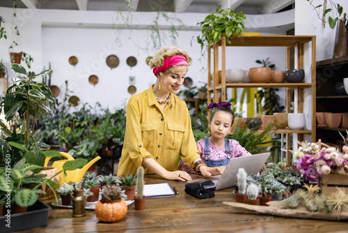 A woman and a girl work together in a flower shop, surrounded by plants and flowers, using a laptop and a notepad